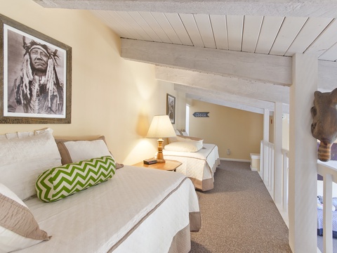 Relax in this peaceful loft bedroom featuring comfortable beds, warm lighting, and unique artwork. Whitewashed wood beams create a cozy, rustic atmosphere perfect for restful nights.