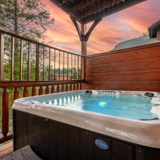 This property's covered deck welcomes a bubbling hot tub surrounded by lush mountain foliage and vibrant sunset skies, creating a peaceful outdoor retreat.