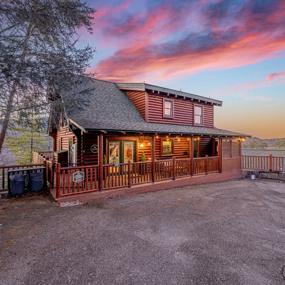 A charming log cabin retreat illuminated against a stunning pink and orange sunset sky, surrounded by peaceful mountain forest.
