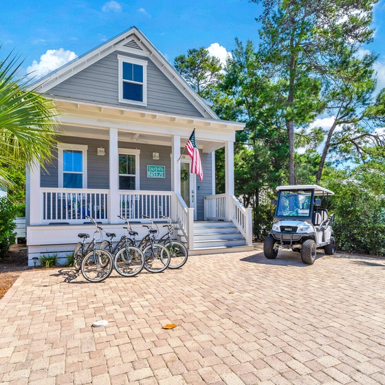 Charming coastal cottage with front porch, bicycles for exploring, and golf cart parking on brick pavers surrounded by lush tropical landscaping.