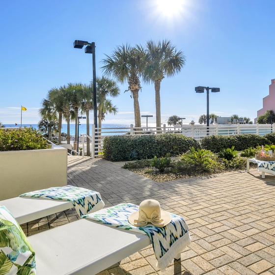 Beachfront ground floor patio featuring loungers with tropical-print towels, landscaped grounds, and palm trees overlooking the water.