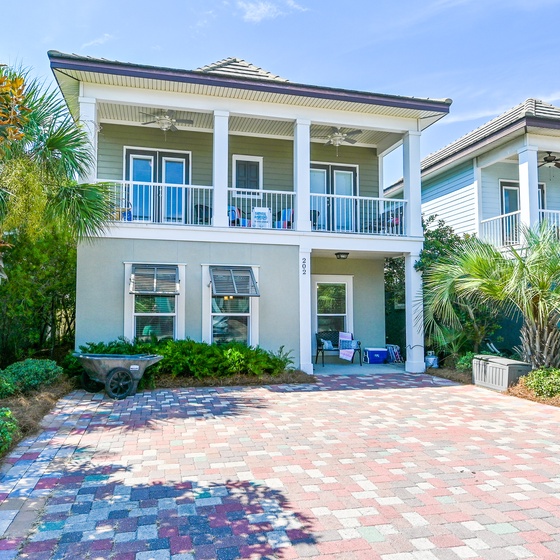 Elegant coastal home with covered porch, tropical landscaping, and decorative brick driveway creates a welcoming arrival experience.