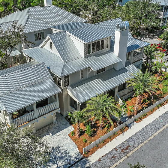 Aerial view of a beautiful coastal home with metal roofing and tropical landscaping in a tree-lined neighborhood.