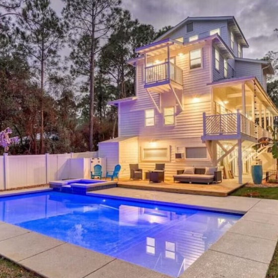 Modern multi-level beach house with illuminated pool area, surrounded by pine trees and featuring multiple decks and balconies.