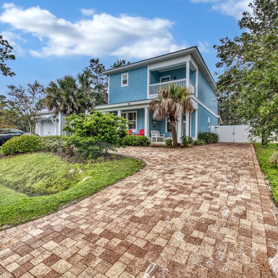 A charming coastal home with bright blue siding welcomes guests, featuring a paver driveway, lush tropical landscaping, and an inviting second-story balcony perfect for relaxing in the sunny Florida weather.