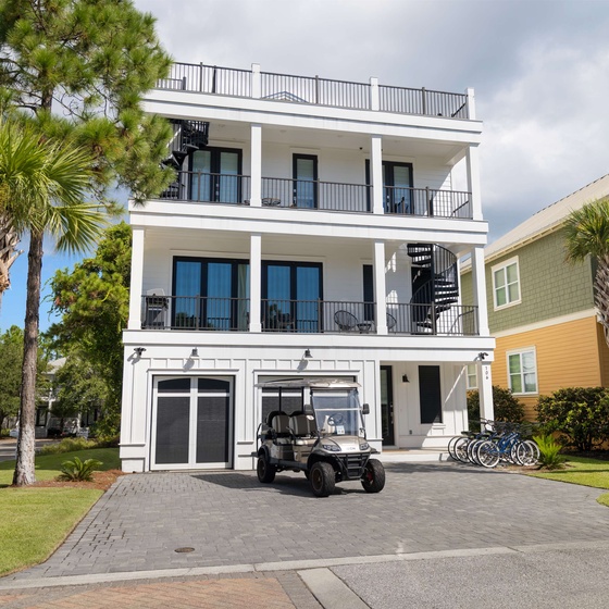 This three-story coastal property features multiple balconies, a rooftop deck, and tropical landscaping. The ground-level garage offers parking and a golf cart, with bikes ready for neighborhood exploration.