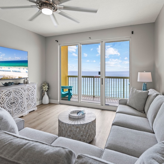 Relax in your coastal living room with ocean views through sliding glass doors, plush gray seating, and beachfront ambiance on the big screen.