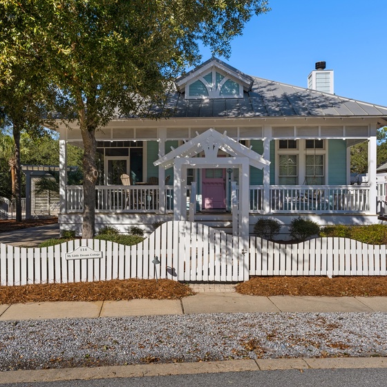 Charming coastal cottage with wraparound porch and picket fence, nestled among mature trees in a peaceful neighborhood setting.