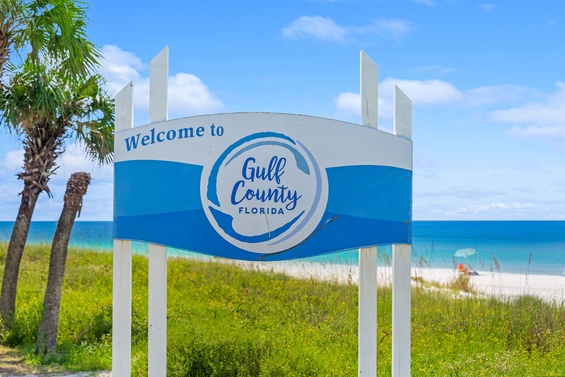 Welcome sign at the entrance to the Gulf County area, featuring white sand beaches and palm trees along the Florida coastline during a sunny day.