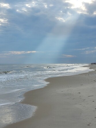 Pristine shoreline stretches beneath dramatic skies where sunbeams pierce through clouds, illuminating gentle waves rolling onto soft sand in this peaceful coastal setting.