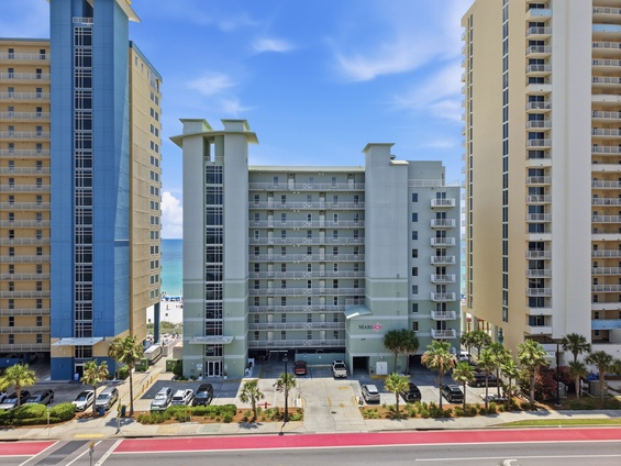 Modern beachfront building nestled among high-rise towers, with palm-lined parking and direct beach access visible in the distance.