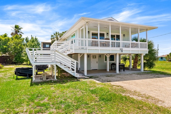 A charming coastal property sits elevated on stilts, featuring a spacious covered porch, outdoor grilling area, and parking underneath, surrounded by tropical palms and blue skies.