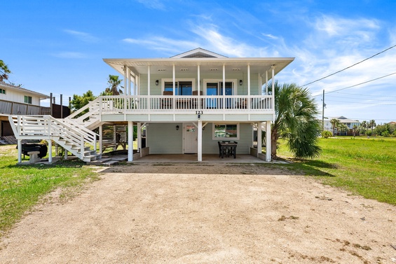 Bright coastal retreat on stilts features a covered ground-level parking area, expansive upper-level deck, and airy covered porch with sweeping views, situated near palm-lined neighborhoods and open green spaces.