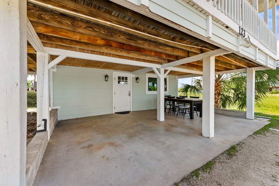 Covered ground-level space beneath the property with parking and an outdoor dining area, offering convenient vehicle access and a shaded spot for gathering near tropical landscaping.