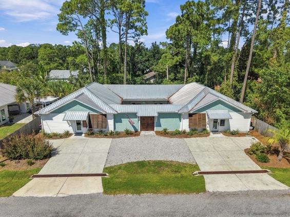 Aerial view of a modern vacation property featuring white and aqua exteriors with crisp metal roofing, nestled among towering pines in a peaceful residential neighborhood.