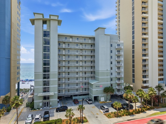 Coastal property building with multiple balconies, situated near the beach with palm trees and parking area at the entrance.