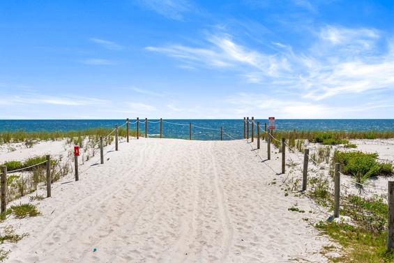 Pristine sandy beach pathway leads directly to sparkling turquoise waters, framed by natural dunes and gentle sea grasses under bright coastal skies.