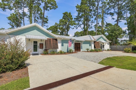A charming coastal-style property nestled among tall pines, featuring soft seafoam green accents and a welcoming front entrance, with ample parking and lush natural landscaping surrounding the home.
