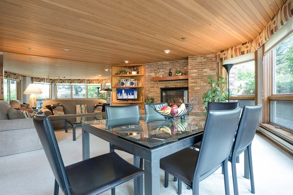Unwind in this warm, open-concept space where natural wood ceilings and brick fireplace create a cozy retreat. Enjoy meals at your dining table while soaking in the tranquil greenery through expansive windows.