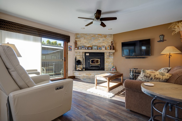 Unwind in your spacious living room, where a stone fireplace adds warmth, plush seating invites relaxation, and natural light streams through sliding doors to the balcony.