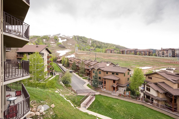 Mountain resort community nestled at the base of ski slopes, featuring residential buildings surrounded by spring greenery and remnants of winter snow on the hillside.