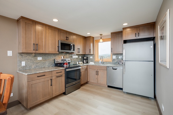 Your fully equipped kitchen features warm wood cabinetry, granite countertops, and modern stainless appliances—perfect for preparing meals while enjoying natural light and mountain vistas through the window.
