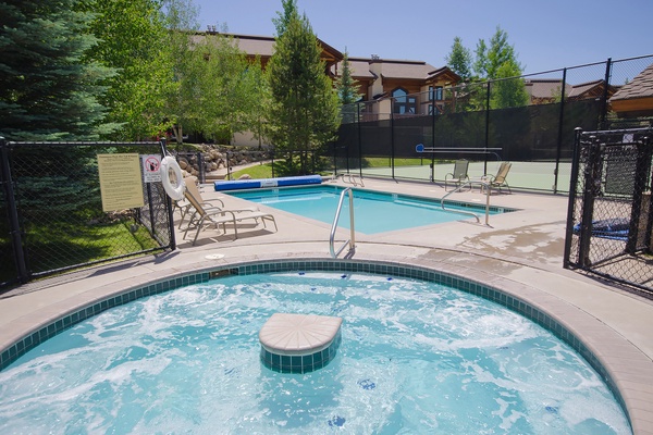 Shared outdoor recreation area featuring heated pool, hot tub, and tennis court surrounded by mature trees and residential buildings in a sunny mountain community setting.