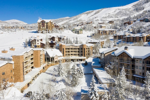 A picturesque alpine village nestled in snow-covered mountains, offering ski-in/ski-out access and a charming mountain retreat. The property sits among scenic slopes and evergreen trees dusted with fresh powder under clear blue skies.