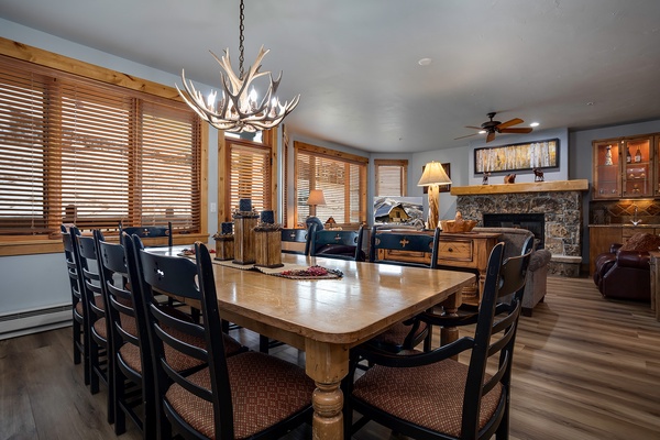 Gather around the expansive dining table under a striking antler chandelier, with mountain-inspired décor and a stone fireplace creating a warm, inviting atmosphere for your group meals.