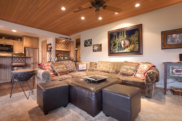 Unwind on the plush sectional beneath warm wood ceilings, where rustic charm meets comfort. Your cozy retreat awaits with inviting seating, nature-inspired art, and an open layout connecting living and dining spaces.