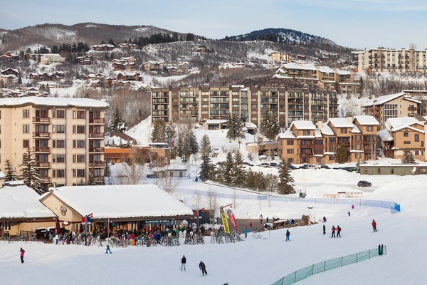 A vibrant winter resort area with ski slopes, lodges, and alpine village buildings nestled among snow-covered mountains and evergreen trees.