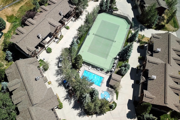Aerial view of a vacation rental community featuring a tennis court, swimming pool, hot tub, and surrounding residential buildings nestled among mature trees and landscaped grounds.