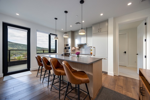 Open-concept kitchen with sleek pendant lights above a spacious island, leather bar stools, and floor-to-ceiling windows framing stunning mountain views.
