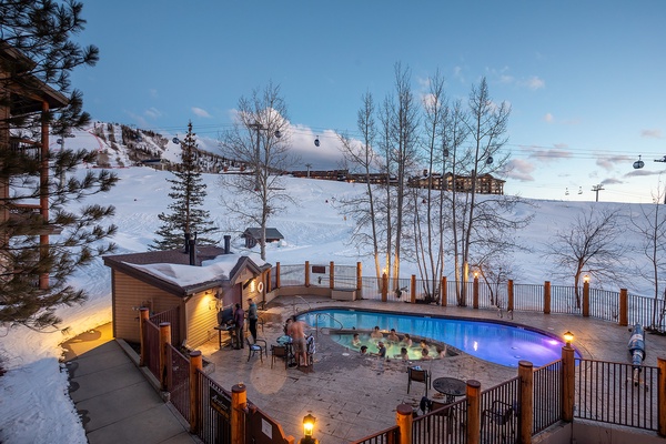 A ski resort featuring an outdoor heated pool surrounded by snow-covered slopes, ski lifts, and rustic lodges at dusk.
