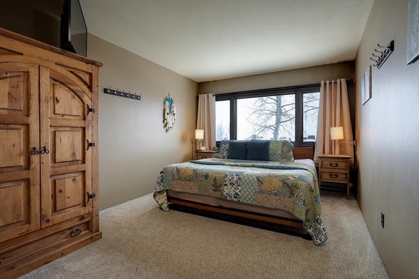 Unwind in your peaceful bedroom with rustic wood accents, plush quilted bedding, and a large window framing serene winter views—your perfect mountain retreat.