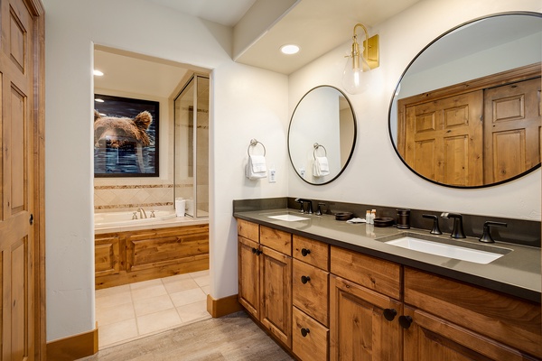Unwind in your private spa-like bathroom with double vanities, elegant round mirrors, and a luxurious soaking tub framed by warm wood cabinetry and charming mountain-inspired artwork.