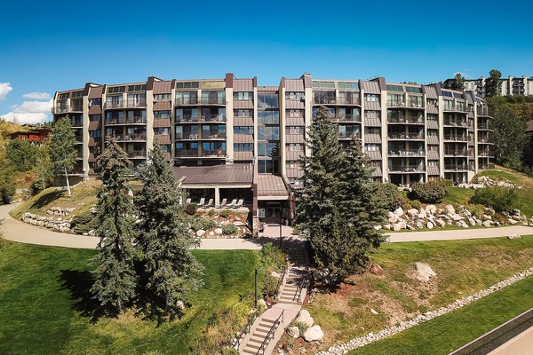 Modern multi-story property nestled in landscaped grounds, surrounded by mature evergreens and natural stone features under a clear blue sky.