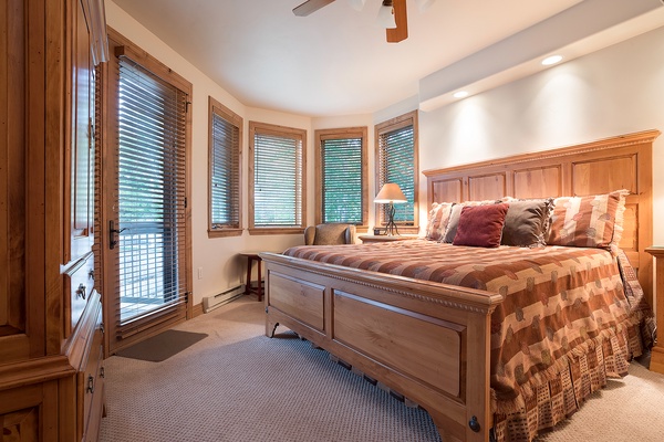 Settle into your cozy bedroom with rich wood furnishings and windows framing peaceful forest views, where soft bedding and gentle lighting invite deep, restful sleep.