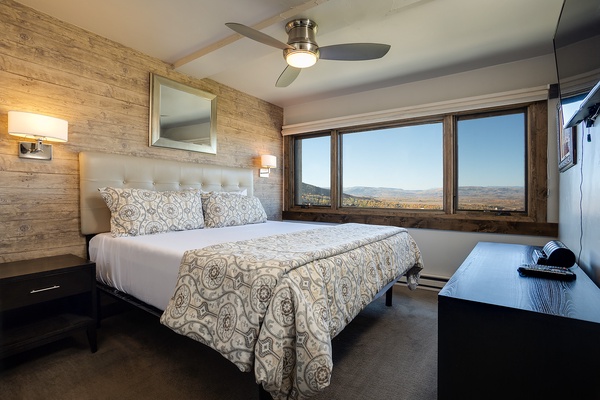 Your serene bedroom features a plush bed with elegant linens, warm wood accents, and stunning panoramic mountain views through expansive windows—perfect for restful mornings.