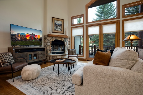 Picture yourself unwinding here—plush seating frames a stone fireplace, while tall windows reveal lush evergreens and a private deck. A cozy space designed for your mountain retreat.