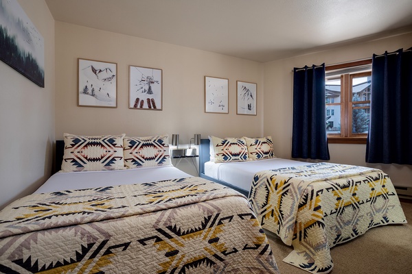 Unwind in your cozy mountain retreat with two comfortable beds dressed in geometric patterns, framed alpine art above, and blackout curtains for restful mornings after adventure-filled days.