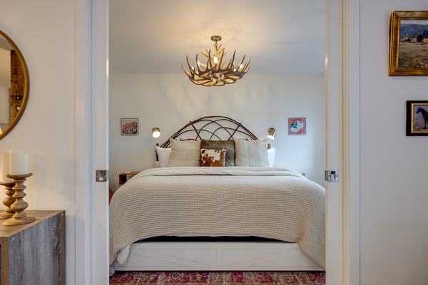 Your bedroom awaits with a plush bed, rustic antler chandelier, and cozy textures. Original artwork and thoughtful touches create a warm, inviting retreat for restful nights.