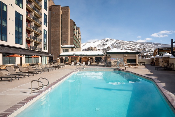 The property features an outdoor pool surrounded by lounge chairs, with stunning snow-covered mountain views framing the ski resort setting.
