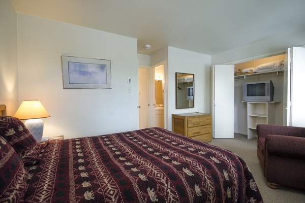 Your cozy bedroom features a plush bed with warm patterned linens, a comfortable armchair, soft lighting, and your own TV. The ensuite bathroom and closet storage keep everything you need close at hand.