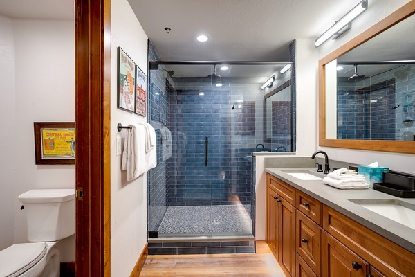 Step into your spa-inspired bathroom featuring a stunning walk-in shower with sleek blue subway tiles, rainfall showerhead, and dual vanity with warm wood cabinetry—perfect for unwinding after your adventures.