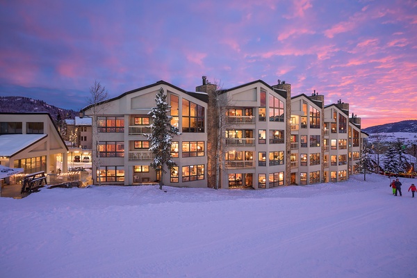Modern mountain property nestled in a snowy alpine setting, illuminated at dusk with striking sunset skies painting the horizon in vibrant pink and purple hues.