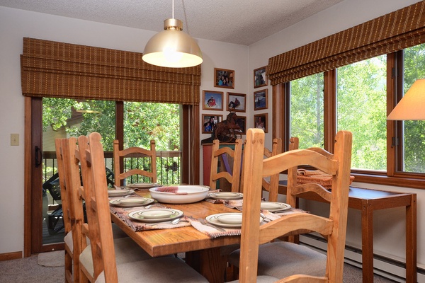 Gather around this warm, inviting dining table where your group can share stories and meals together, surrounded by natural light streaming through large windows that frame lush green views.