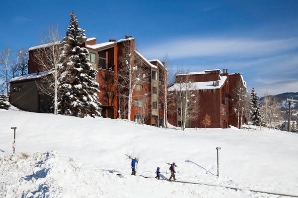 Mountain vacation rental complex featuring rustic wood and stone architecture nestled in a snowy alpine setting, with ski-in access and surrounded by evergreens.