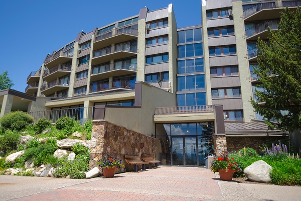 A modern multi-story property with multiple balconies, surrounded by landscaped gardens featuring natural stone accents and colorful seasonal flowers, offering comfortable accommodation under clear blue skies.