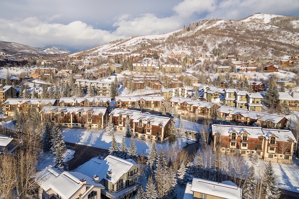 Aerial perspective of a snow-covered mountain resort community, showcasing vacation rental properties nestled among evergreen trees with ski slopes and peaks rising in the distance.